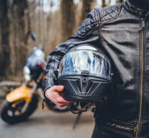 Person in leather jacket holding their helmet in front of motorcycle.
